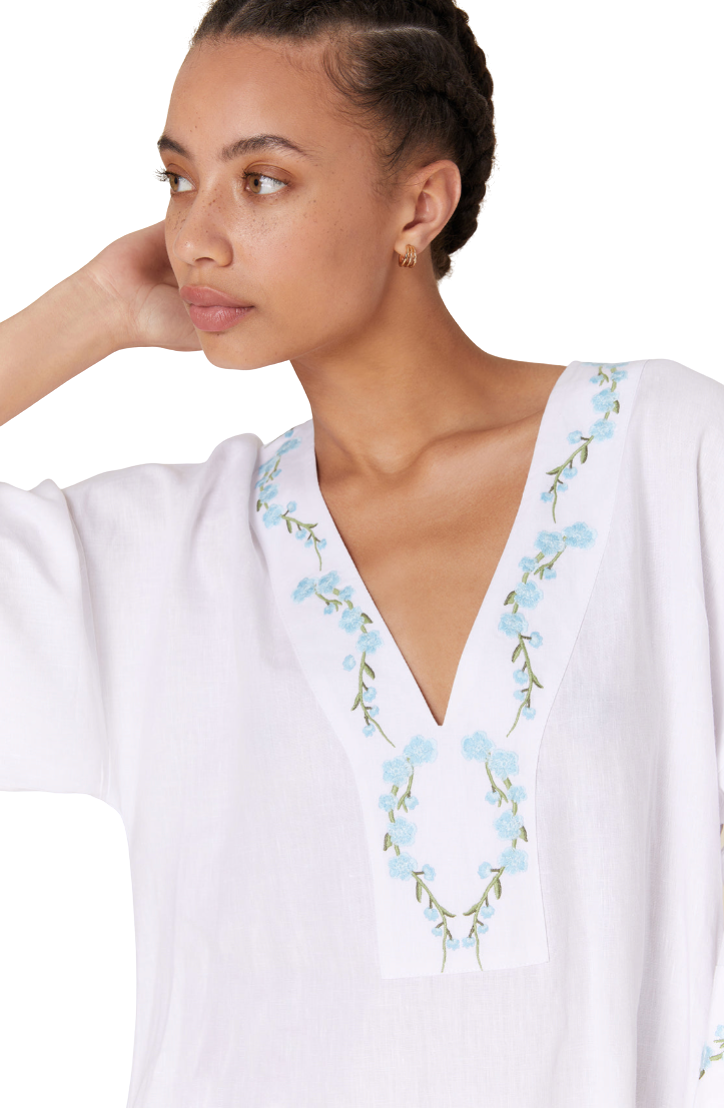 A woman wearing a white V-neck tunic top with light blue and green floral embroidery around the neckline, shown against a plain white background.