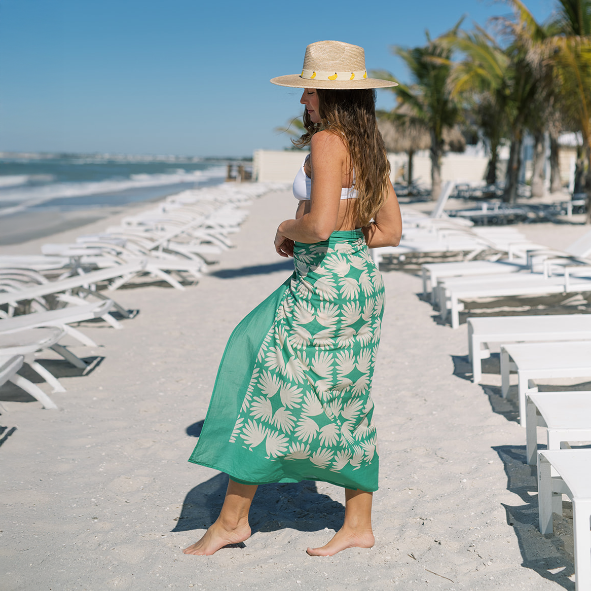 Woman in a green patterned sarong and straw hat on a beach.