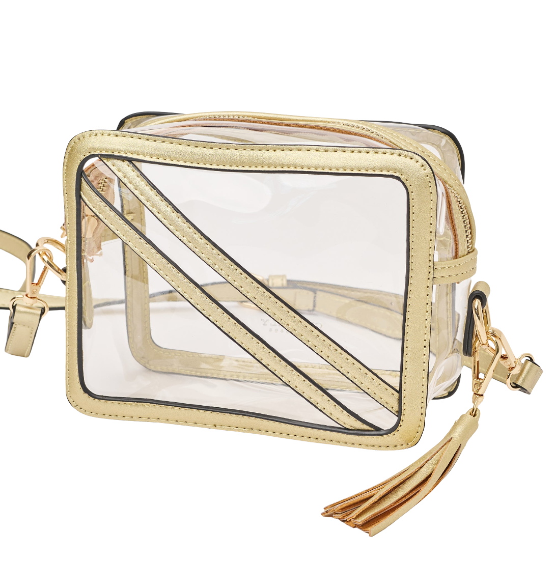 A clear plastic cosmetic bag with gold-tone trim, zipper, and strap, featuring diagonal gold straps across the front and a matching tassel charm, shown on a plain white background.