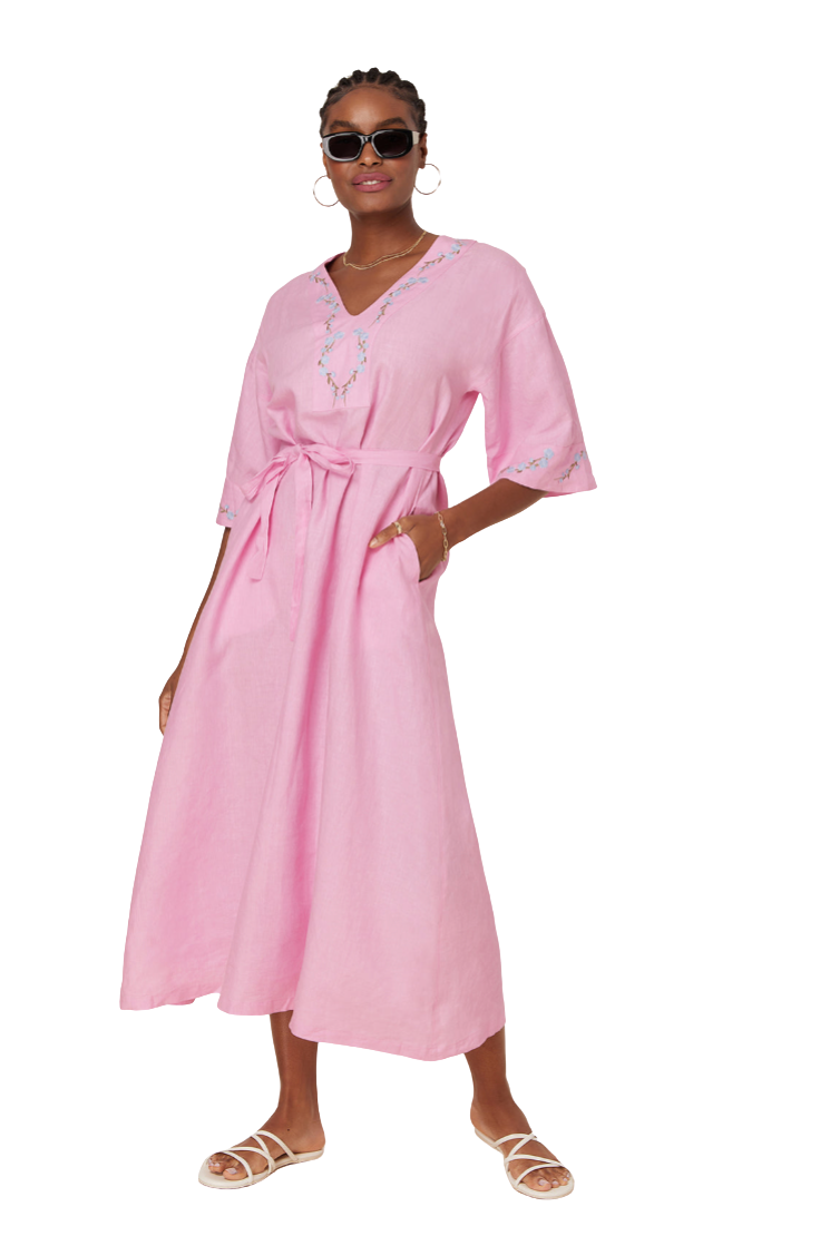 A woman modeling a light pink kaftan dress with embroidered details on the neckline and sleeve cuffs, paired with white strappy sandals and sunglasses, set against a plain white background.