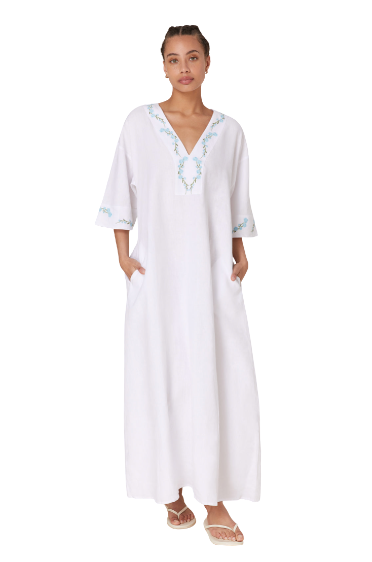 A woman modeling a white kaftan dress with light blue and green embroidered details on the neckline and sleeve cuffs, paired with neutral sandals, set against a plain white background.