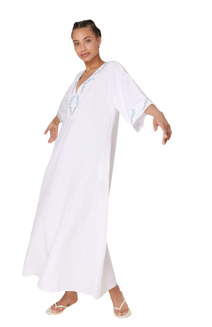 A woman modeling a white kaftan dress with light blue embroidered details on the neckline and sleeve cuffs, set against a plain white background.