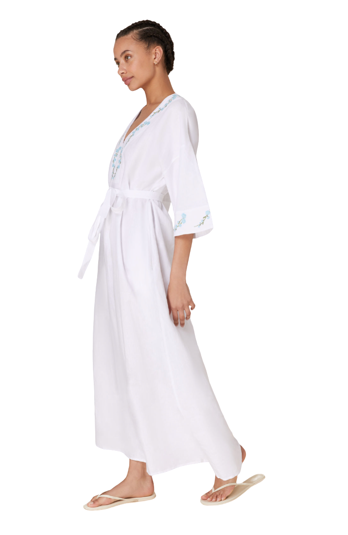 A woman modeling a white maxi robe with light blue floral embroidery on the neckline and sleeve cuffs, paired with beige flip-flop sandals against a plain white background.