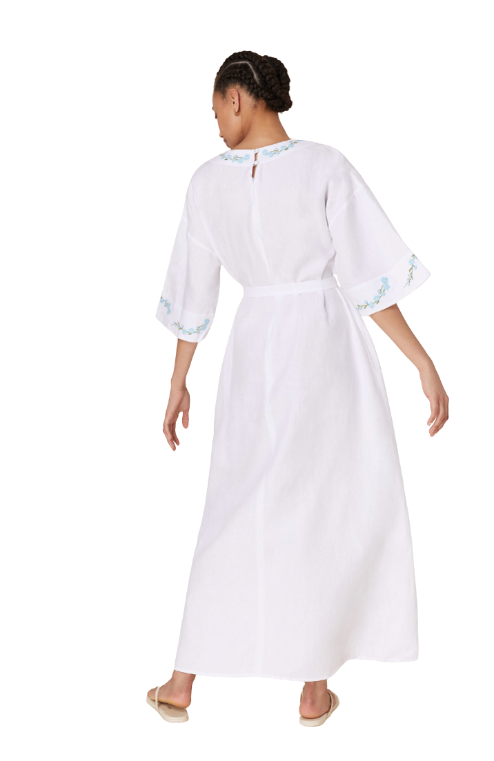 A woman shown from the back wearing a white maxi dress with light blue floral embroidery on the collar and sleeve cuffs, paired with white slide sandals, against a plain white background.
