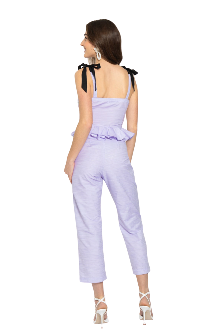 A woman modeling a light purple cropped jumpsuit with black ribbon shoulder ties, shown from the back against a plain white background.