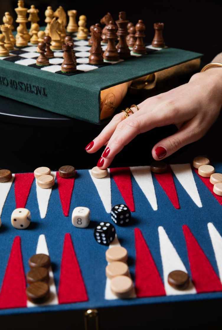 A close-up lifestyle shot of a hand with red nail polish reaching for a backgammon piece on a blue, red, and white felt board. In the background, a person is seen playing with a wooden chess set on a green book.