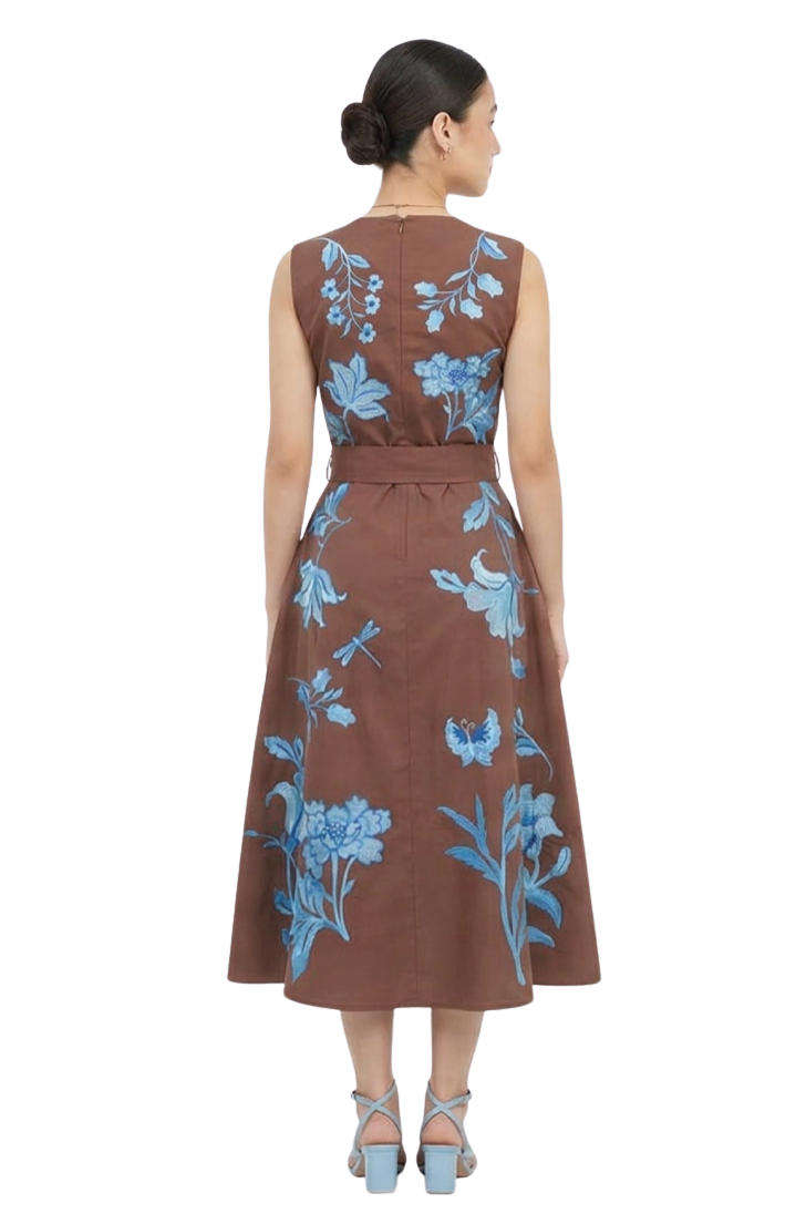 A woman shown from the back wearing a sleeveless brown midi dress with a blue floral and dragonfly print, paired with light blue heeled sandals against a plain white background.