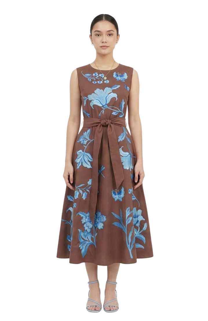 A woman modeling a sleeveless brown midi dress with a blue floral and butterfly pattern, featuring a tie waist and A-line skirt, set against a plain white studio background.