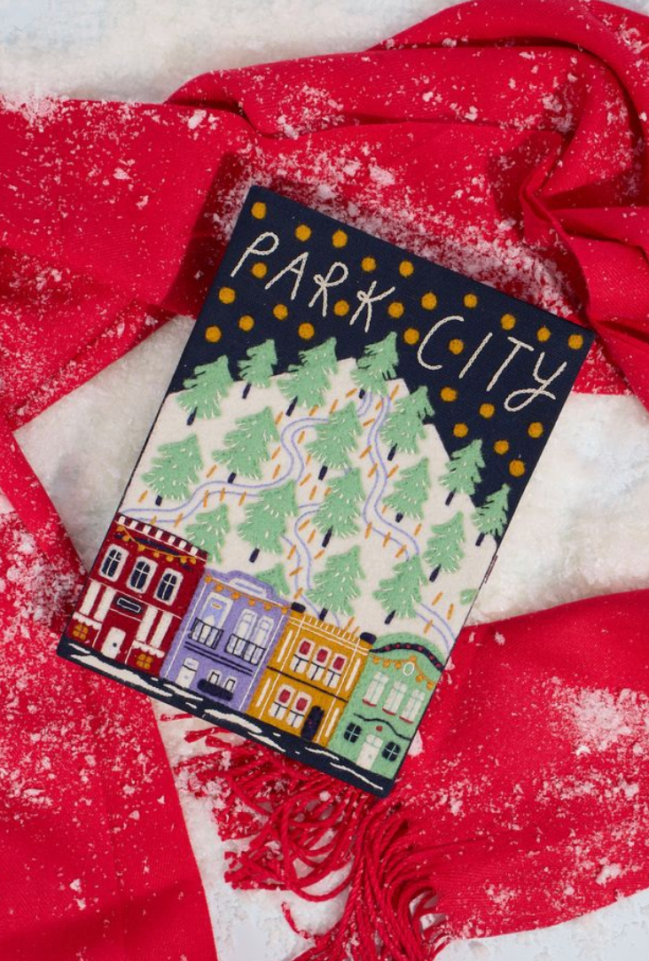 A square art print or book cover titled 'PARK CITY' featuring a winter town illustration with colorful houses, green trees, winding roads, and gold dots on a dark blue background, styled on red and white fabric with white speckles.