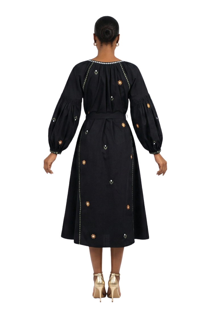 A woman viewed from the back wearing a black midi dress with long balloon sleeves, a self-tie waist belt, and small embroidered floral details. She is standing against a plain white studio background.