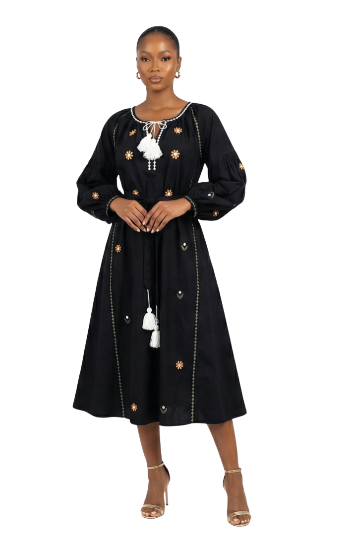 A woman modeling a black midi dress with long sleeves, embroidered details, and white tassels, photographed against a plain white studio background.