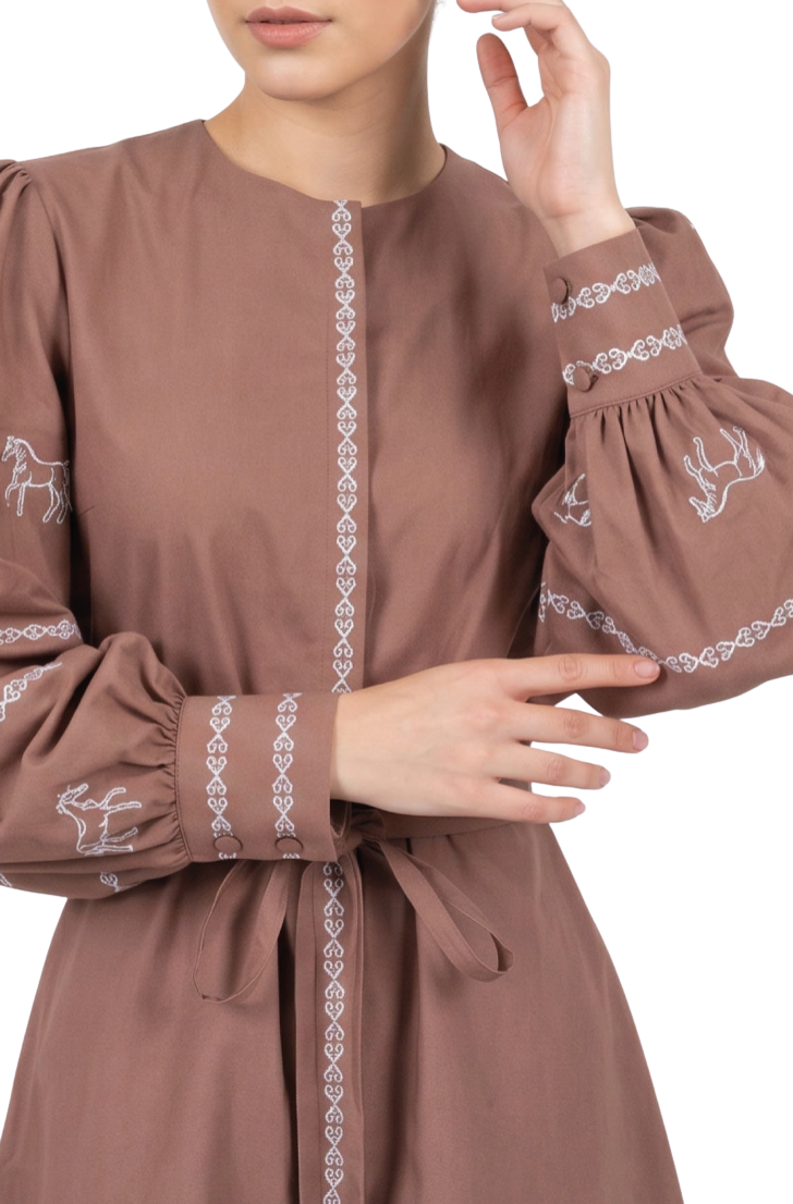 A close-up of a woman wearing a brown long-sleeve dress with white embroidered details, including a vertical pattern down the front and motifs on the sleeves.