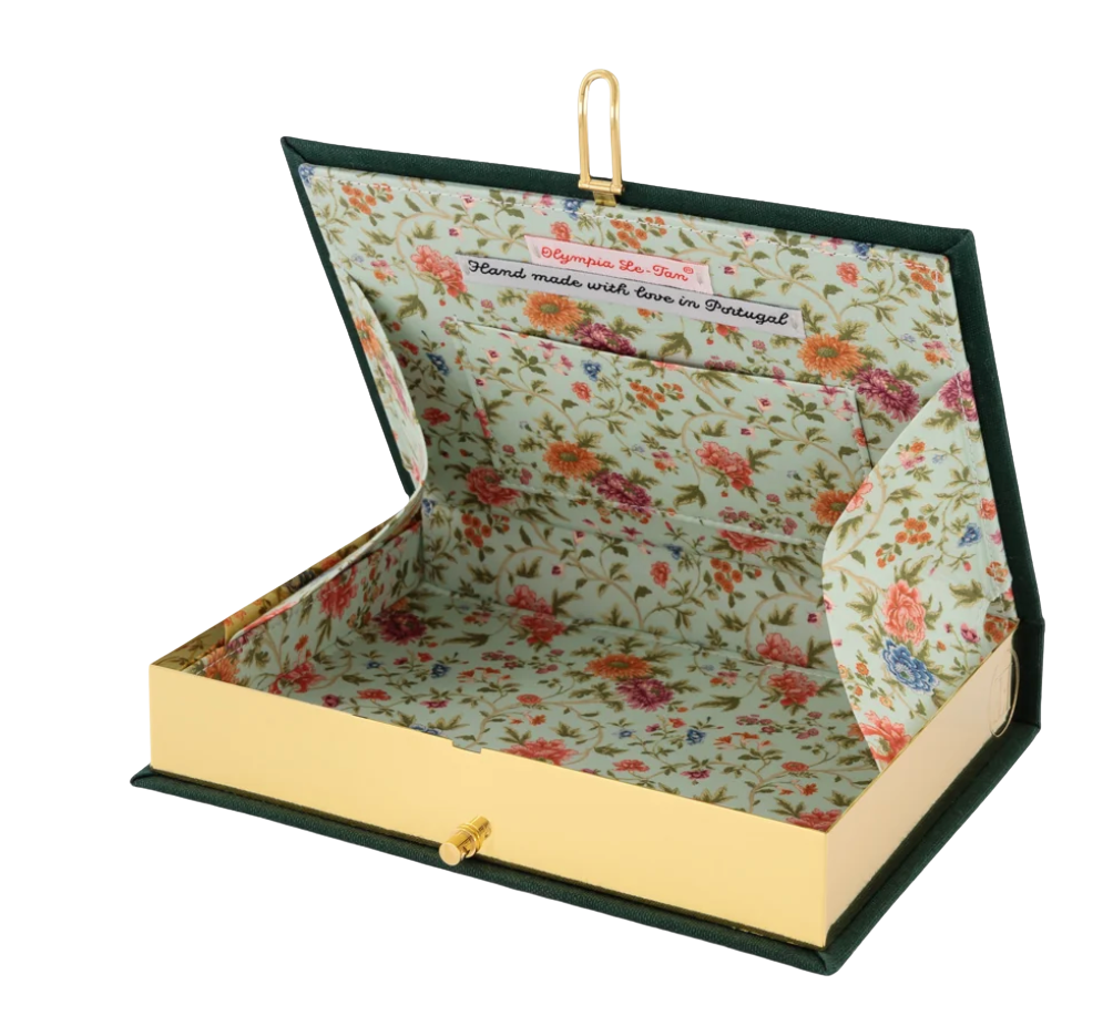 A book-shaped jewelry box with a dark green cover, gold-tone clasp and front pull, and a floral-patterned interior. The lid is open, showing a floral print with pink, orange, and blue flowers on a light background, with a small hanging loop at the top.