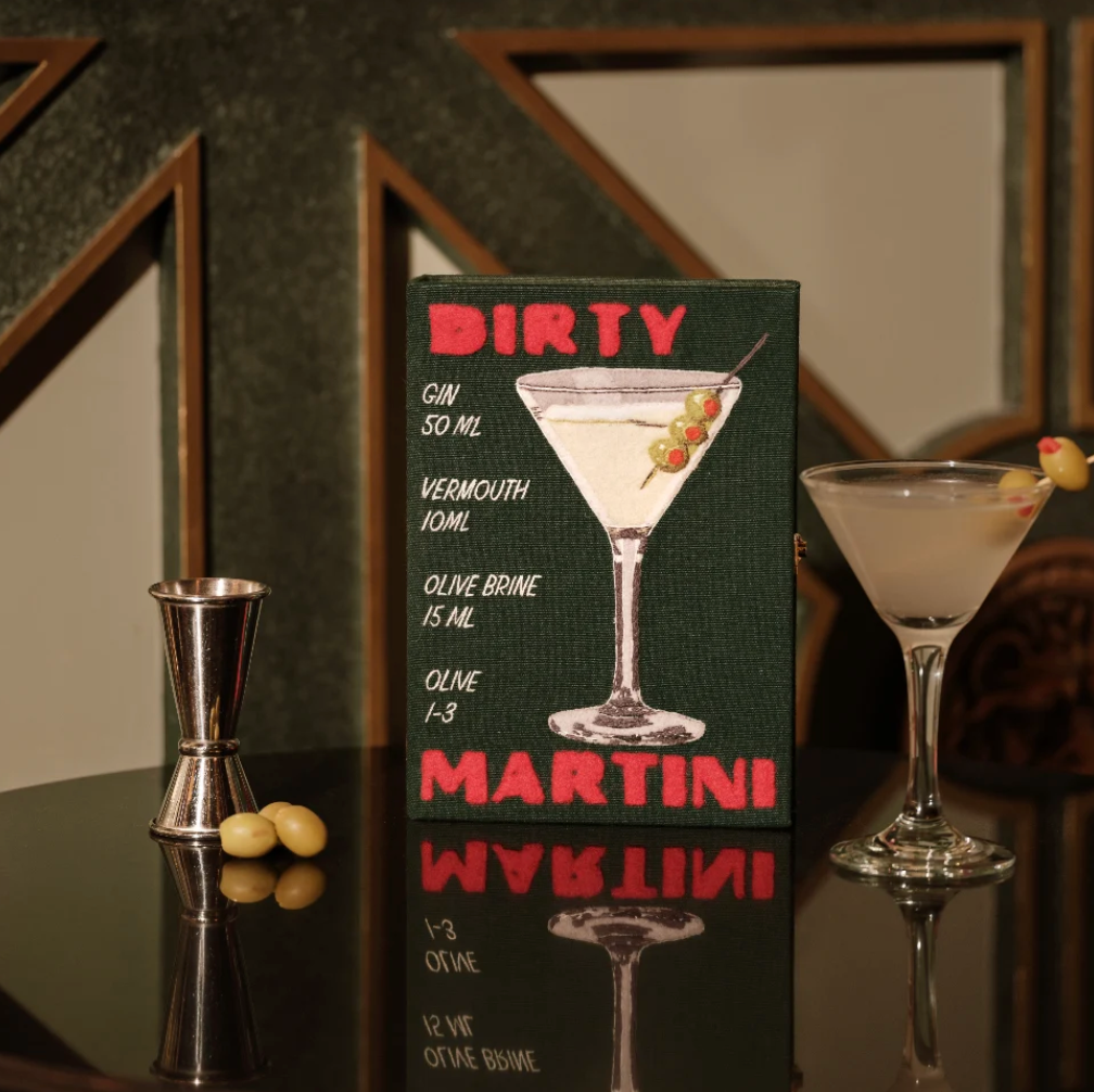 A rectangular cocktail sign reading 'DIRTY MARTINI' with a martini glass illustration and ingredients listed, standing on a dark reflective surface beside a metal jigger, olives, and a martini glass.