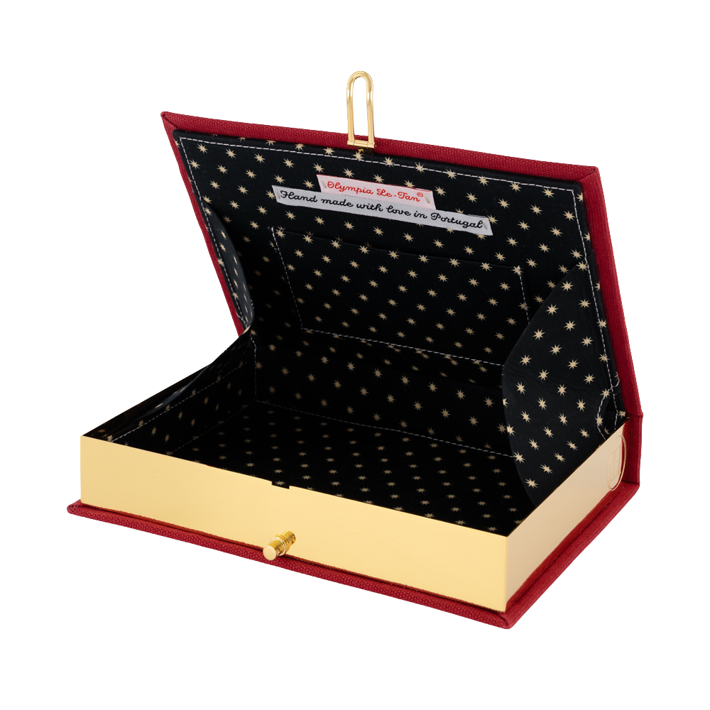 An open book-shaped jewelry box with a red velvet exterior, gold-tone metal trim and clasp, and a black interior lined with gold stars. A small white tag is visible inside the lid.