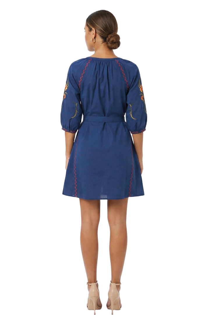 A woman viewed from the back wearing a navy blue dress with three-quarter puff sleeves, a tie waist, and red and yellow embroidered details on the sleeves and sides, paired with beige high-heeled sandals against a white studio background.