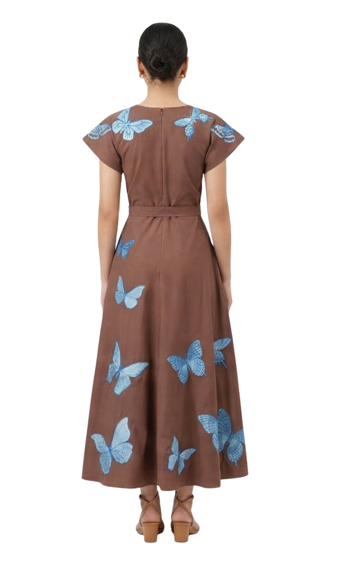 A woman viewed from the back wearing a brown midi dress with blue butterfly prints, paired with brown heeled sandals, against a plain white background.
