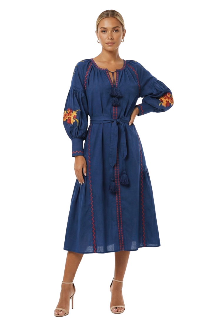 A woman modeling a navy blue midi dress with red embroidered details, tassel ties, and yellow floral embroidery on the sleeves, set against a plain white studio background.