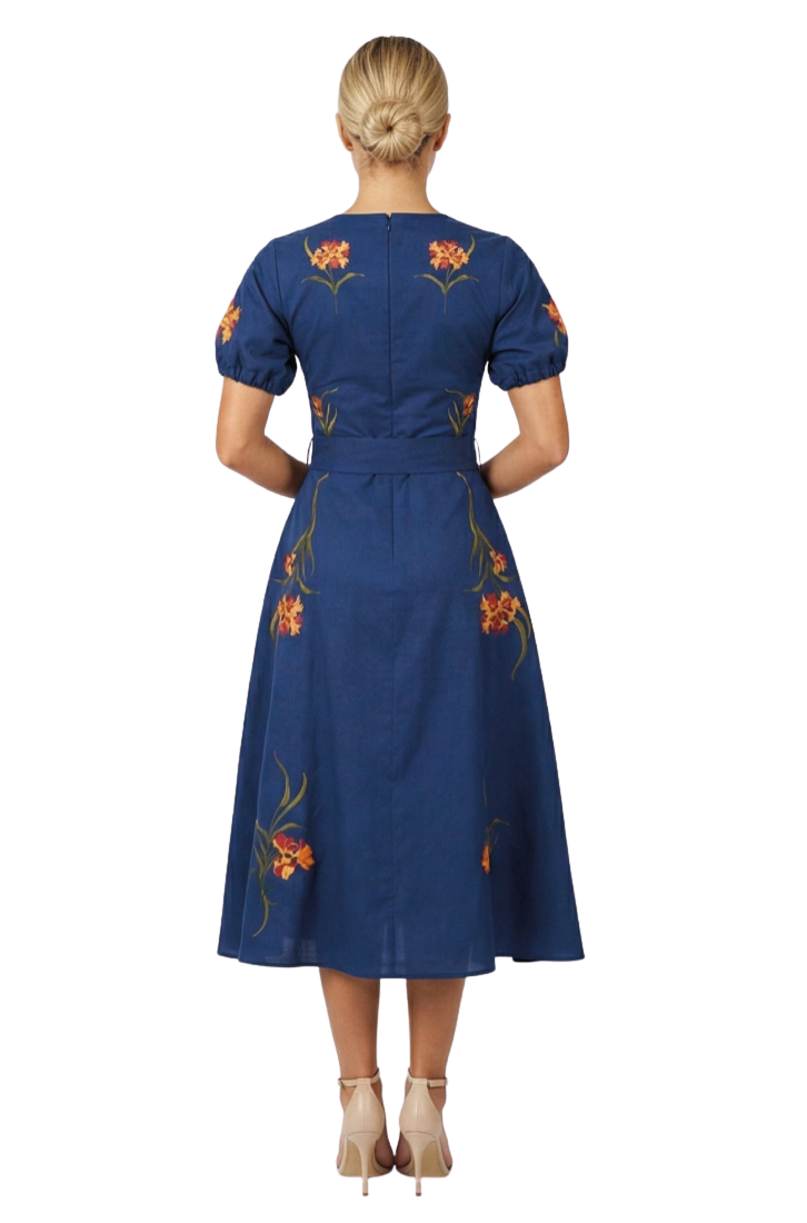 A woman viewed from the back wearing a navy blue midi dress with short puff sleeves, a matching waist belt, and orange floral embroidery with green leaves. She is standing against a plain white studio background.