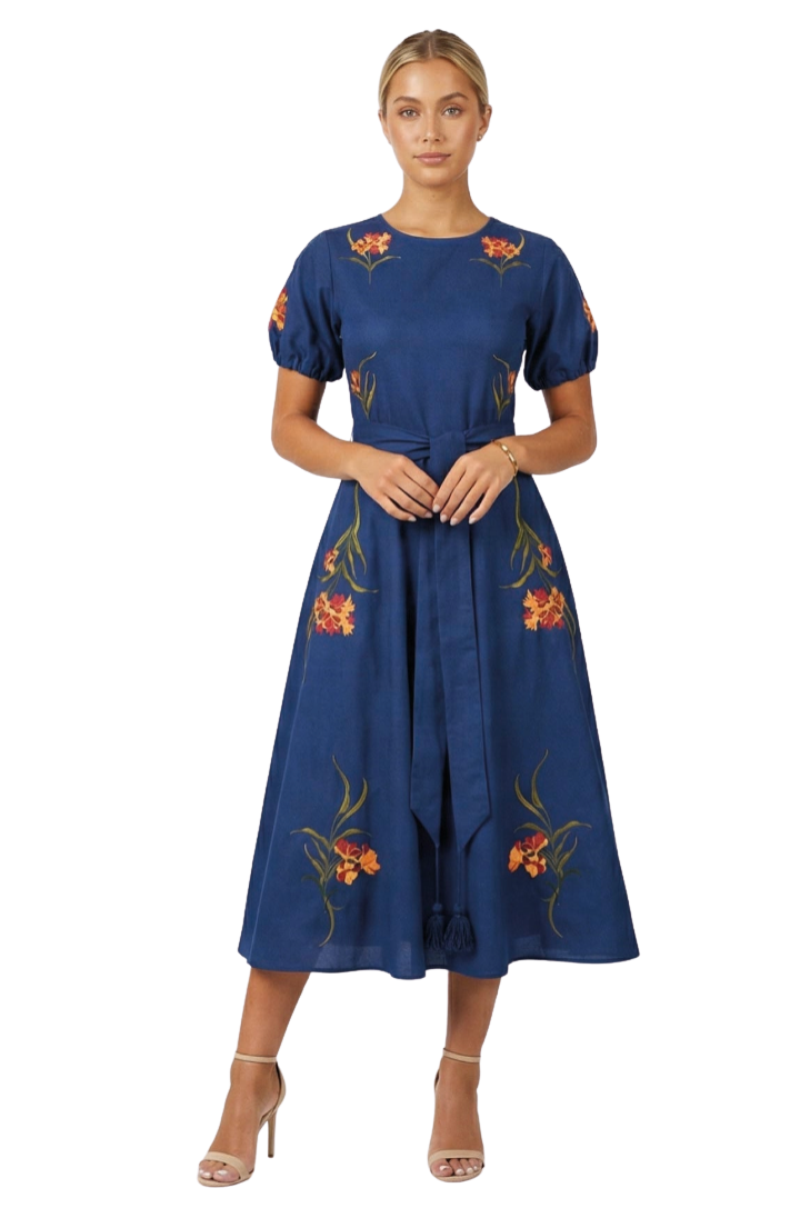 A woman modeling a navy blue midi dress with short puff sleeves and orange floral embroidery, standing against a plain white studio background.