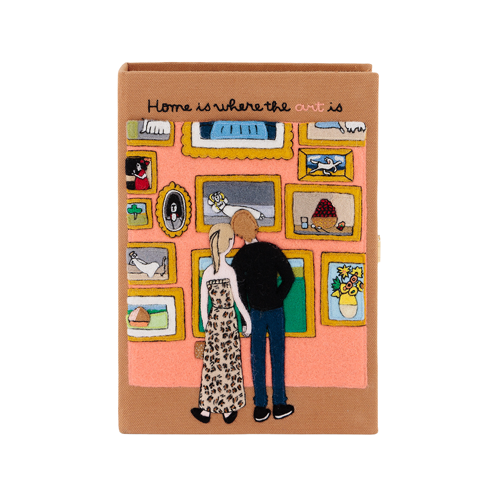 A rectangular book or notebook with a tan cover, featuring an illustration of a couple from behind looking at framed art on a pink wall. The cover has the text 'Home is where the art is' written across the top.