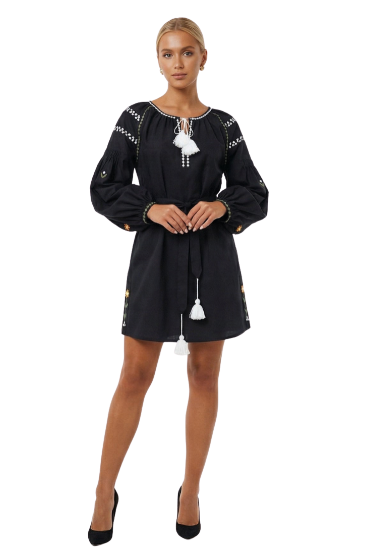 A woman modeling a black short dress with white embroidered details, tassel ties, and white geometric patterns on the shoulders, paired with black high heels against a white studio background.