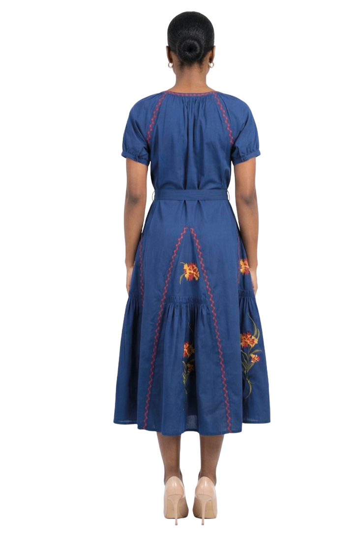 A woman viewed from the back wearing a blue midi dress with red embroidered stitching and floral details, paired with beige high heels, against a plain white background.