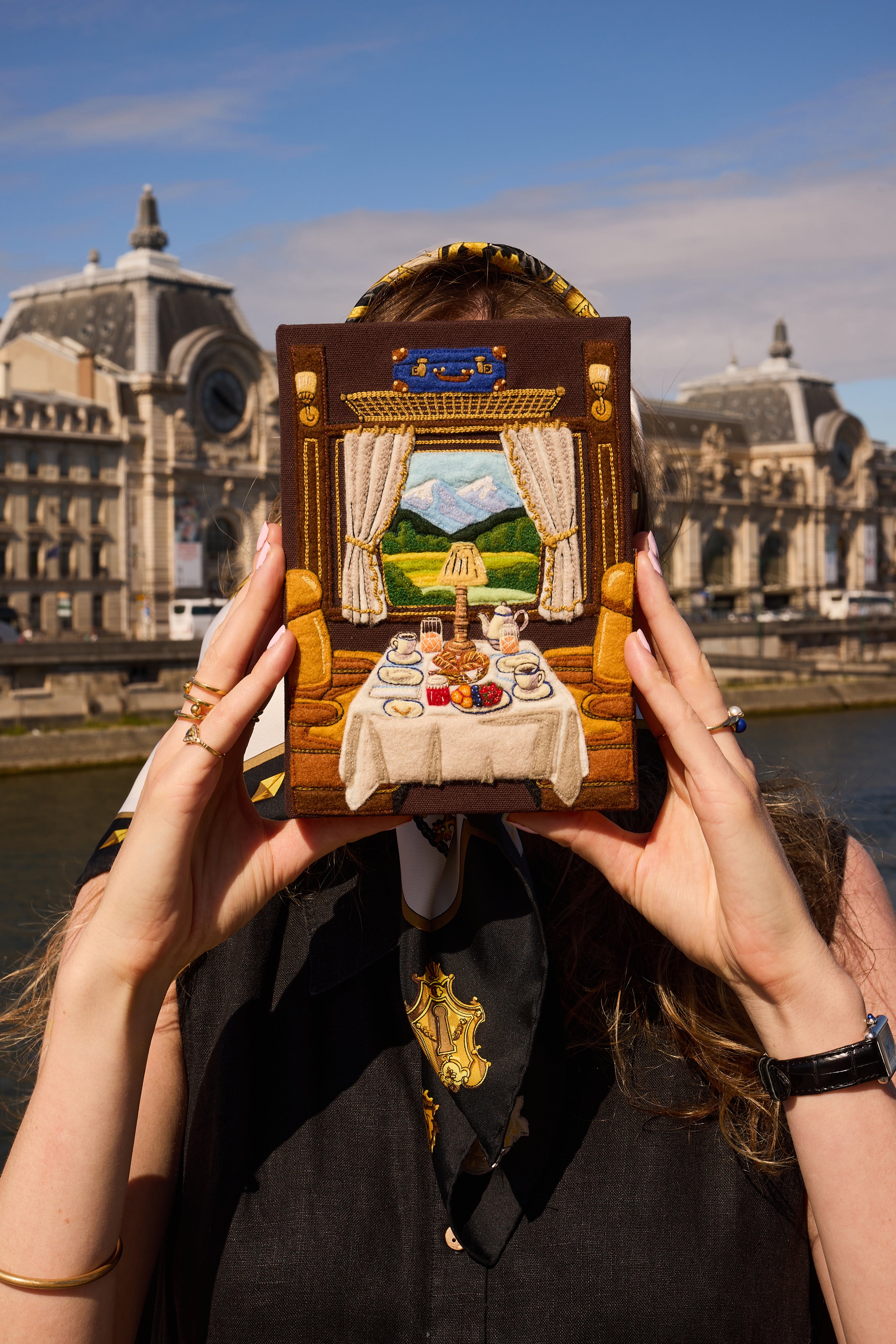 A person outdoors holding a miniature diorama model of a dining room scene in front of their face, with a large historic building and river in the background.