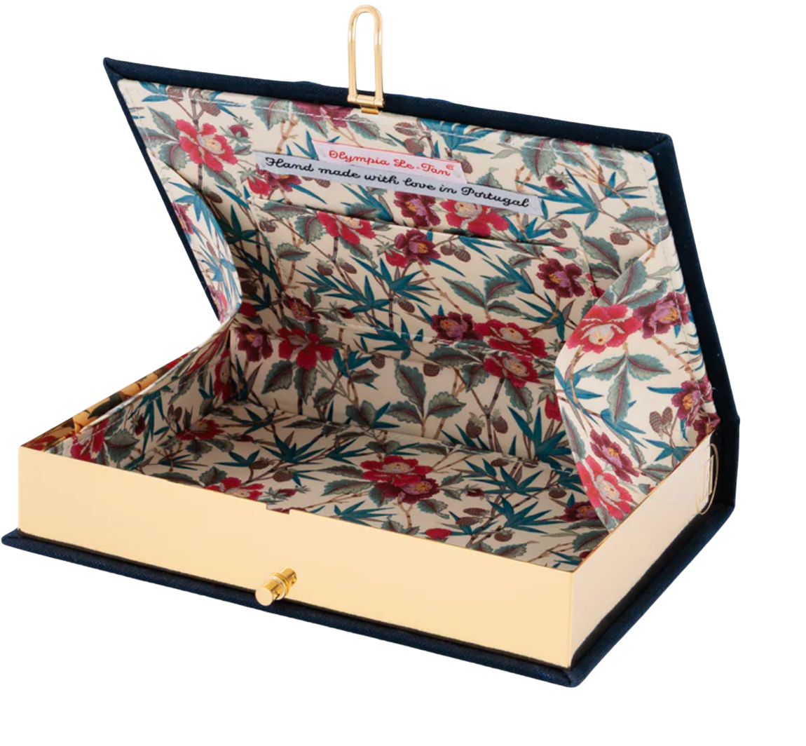 A book-style clutch case with a navy blue velvet exterior, gold-tone hardware, and a floral-patterned interior. The lid is open, showing a cream background with red and green botanical print and a gold clasp.