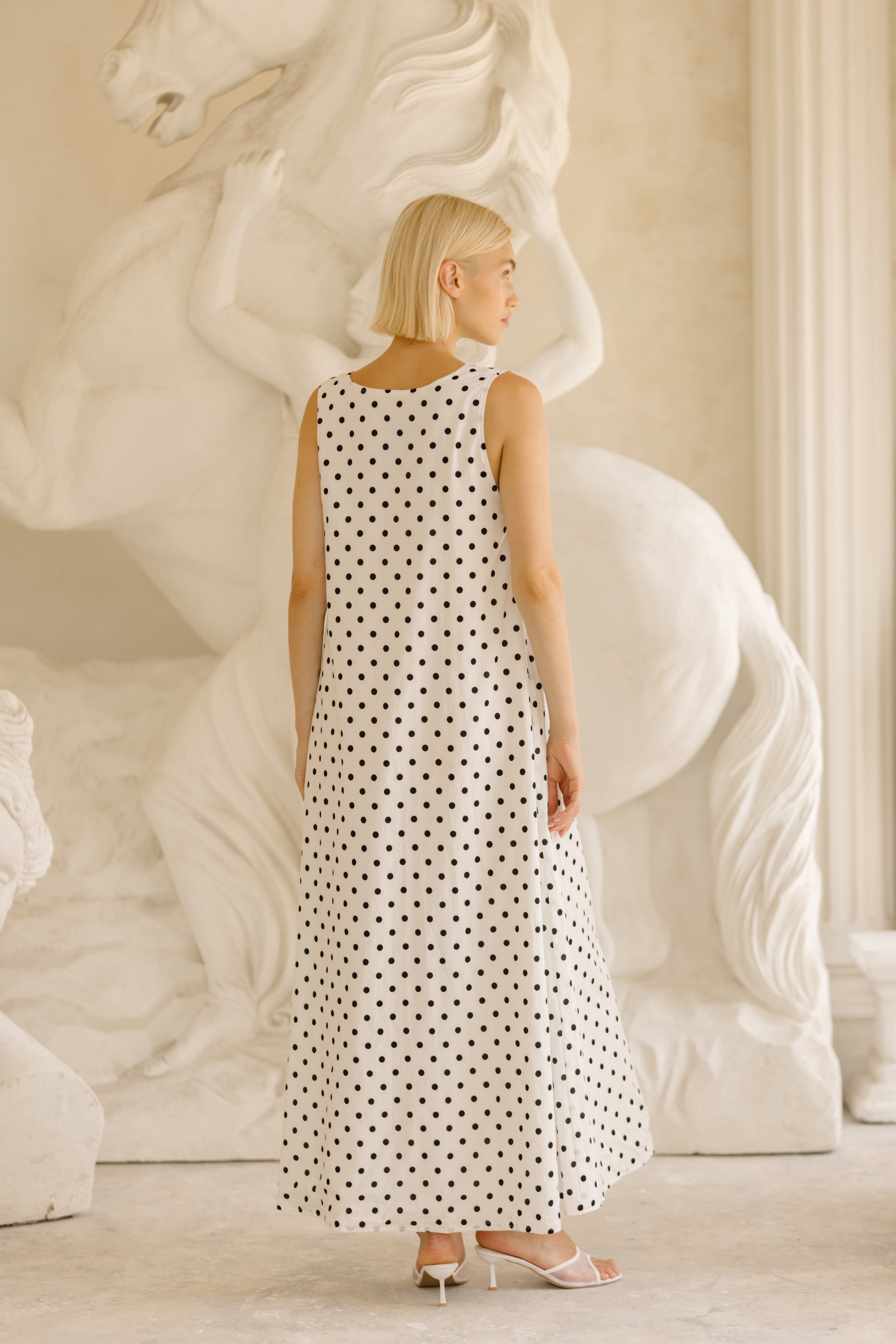 A woman stands in profile wearing a sleeveless white maxi dress with a black polka dot pattern, posed in front of a large white classical horse statue in a bright, elegant interior.